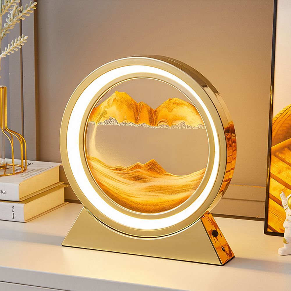 The image shows a white dresser with drawers, books, a flower vase, and a photo frame on top. In the center of the dresser is a gold-colored table lamp with a picture of a mountain scene on it, and in the background is a wall.