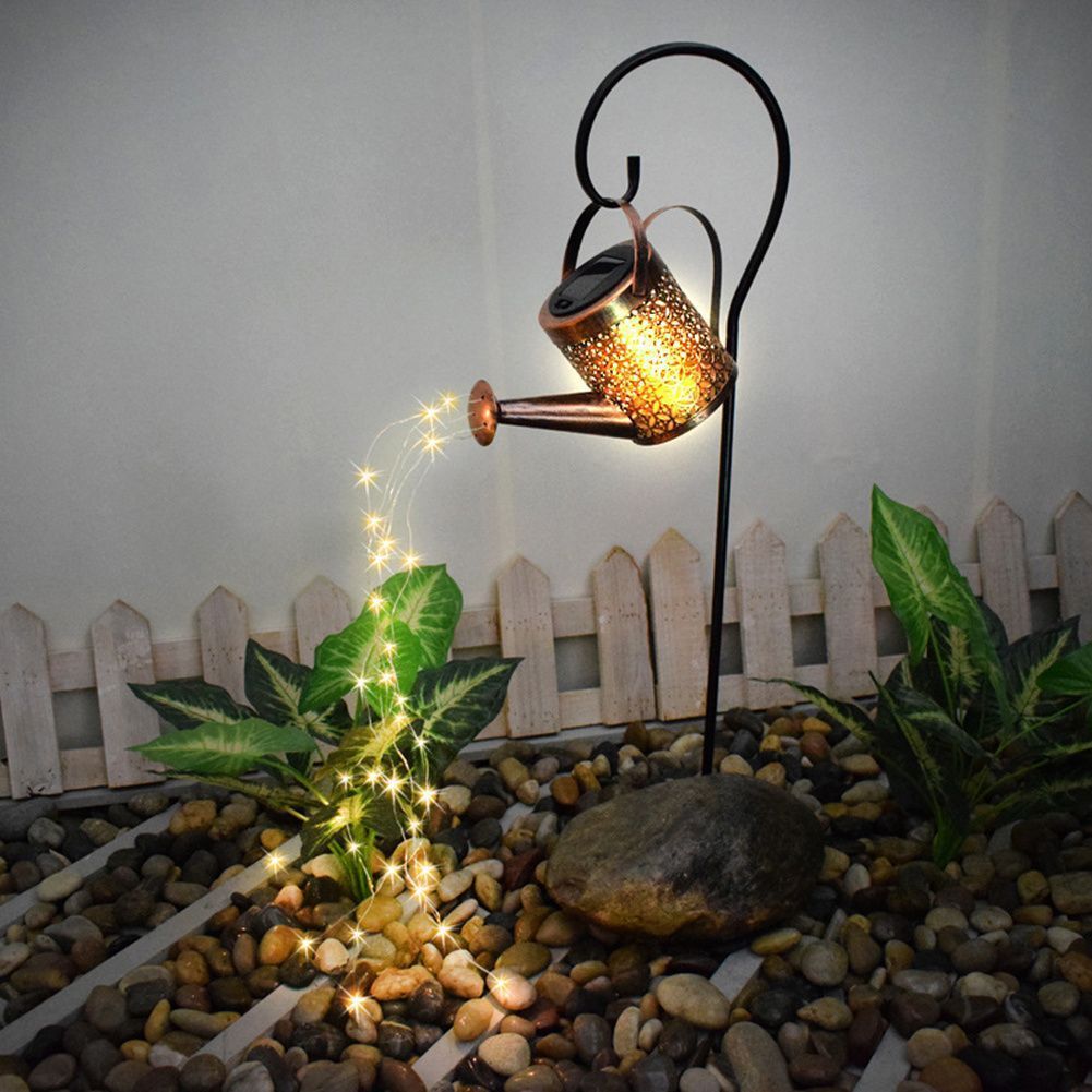 The image shows a solar powered watering can with LED lights in the middle of a garden, surrounded by stones and plants, with a wooden fence in the background and a wall behind it.