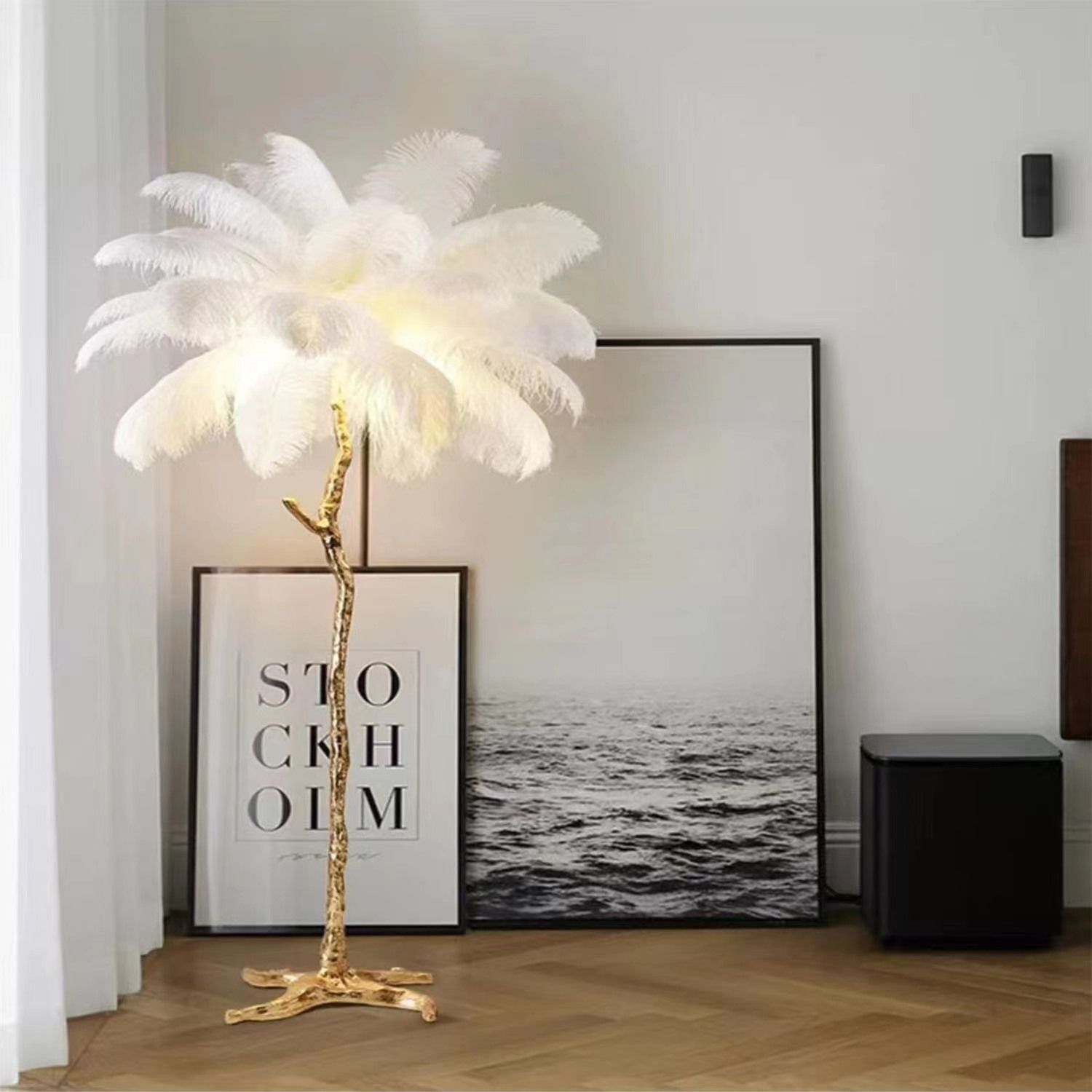 The image shows a white ostrich feather floor lamp in a living room, with a photo frame on the floor, a black object on the right side, and a white wall in the background. The lamp is adorned with white feathers, giving it a luxurious and elegant look.