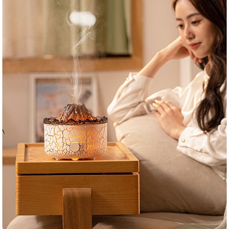 The image shows a woman sitting on a couch next to a wooden table with a humidifier on it. She is holding a cup in her hand and there is a pillow behind her. In the background, there are photo frames on the wall and a window with a curtain. The humidifier is an essential oil diffuser, providing a calming atmosphere to the room.