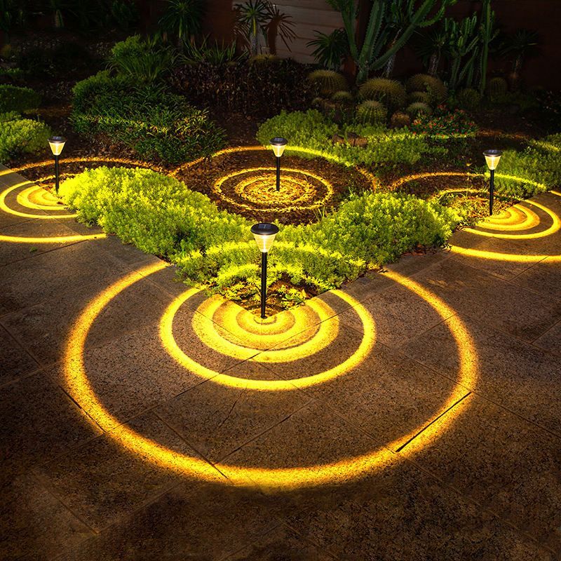 The image shows a garden path illuminated by solar lights in the middle of it at night. The path is surrounded by lush plants and trees, creating a peaceful atmosphere. The lights are arranged in a circular pattern, adding a decorative touch to the garden.