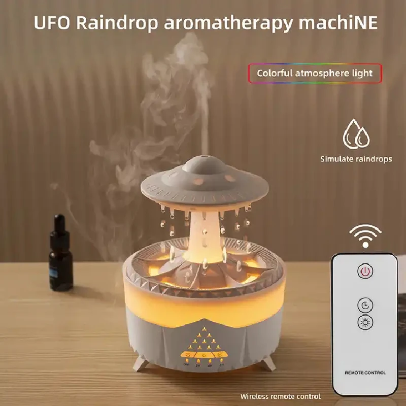 The image shows a UFO raindrop aromatherapy machine on a table with a remote control beside it. The text on the image reads "UFO Raindrop Aromatherapy Machine" and there is a wall in the background.