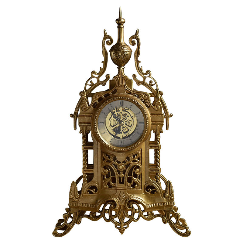 The image is of an antique clock with an ornate design. The clock is made of brass and has a round face with a gold-colored clock face in the center. The face of the clock is decorated with intricate patterns and designs, including swirls, dots, and lines. Above the clock face, there is a tall, ornate finial with a pointed top. The finial is attached to the top of the watch face with two curved arms. The watch face is surrounded by a decorative border with a floral pattern. The overall design is ornate and ornate, with a vintage or antique feel.