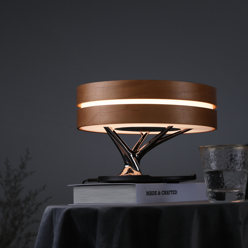 The image shows a modern table lamp with a unique design. The lamp has a round, copper-colored shade that is turned on, creating a warm glow. The shade is made of a shiny, metallic material that reflects the light. The base of the lamp is made up of a series of curved lines that form a tree-like shape, with a black base and a gold-colored base. On the table, there is a stack of books and a glass of water. The background is a dark grey wall, and the overall mood of the image is minimalistic and contemporary.