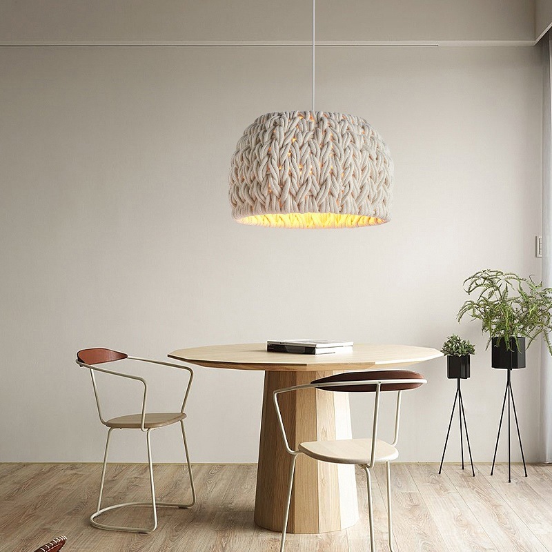 The image shows a modern dining room with a round wooden table and two chairs. The table is in the center of the room and has a book and a plant on it. Above the table, there is a large pendant light with a unique design. The light fixture is made of a woven fabric with a yellow and white color scheme. The walls are painted white and there are two potted plants on either side of the table. The floor is made up of light-colored wood planks. The overall aesthetic of the space is minimal and contemporary.