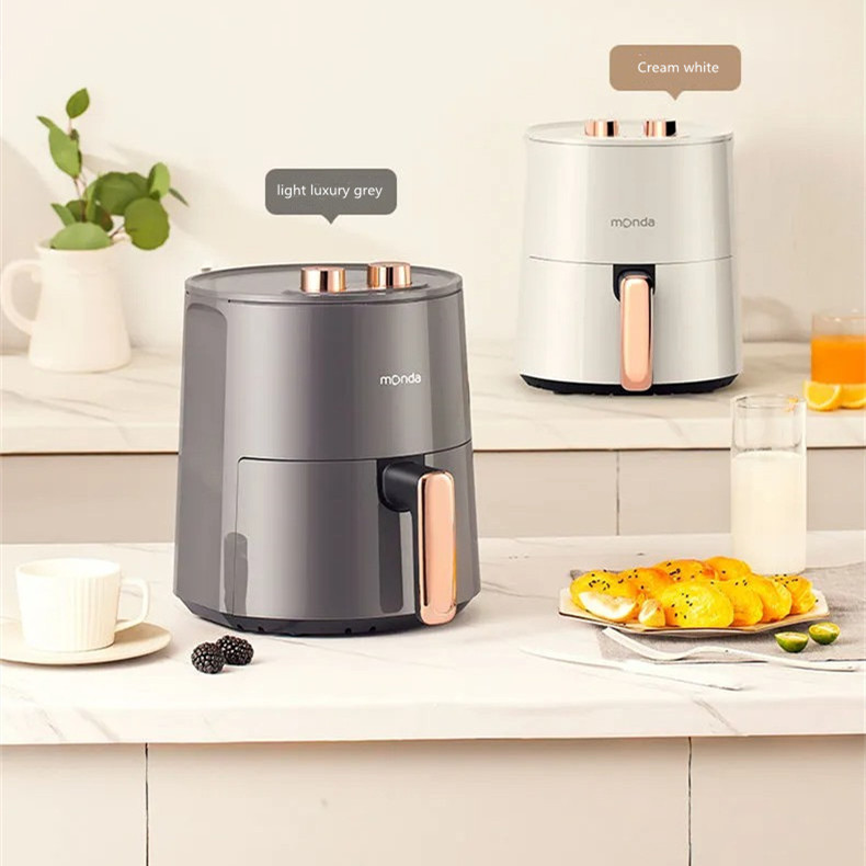 The image shows a kitchen countertop with two modern kitchen appliances. On the left side of the countertop, there is a gray electric juicer with a copper handle and a white cup and saucer. Next to the juicer, there are a few blackberries and a plate of lemons. In the center of the image, there appears to be a glass of orange juice and a small plant in a white vase. The background is a white tiled wall.