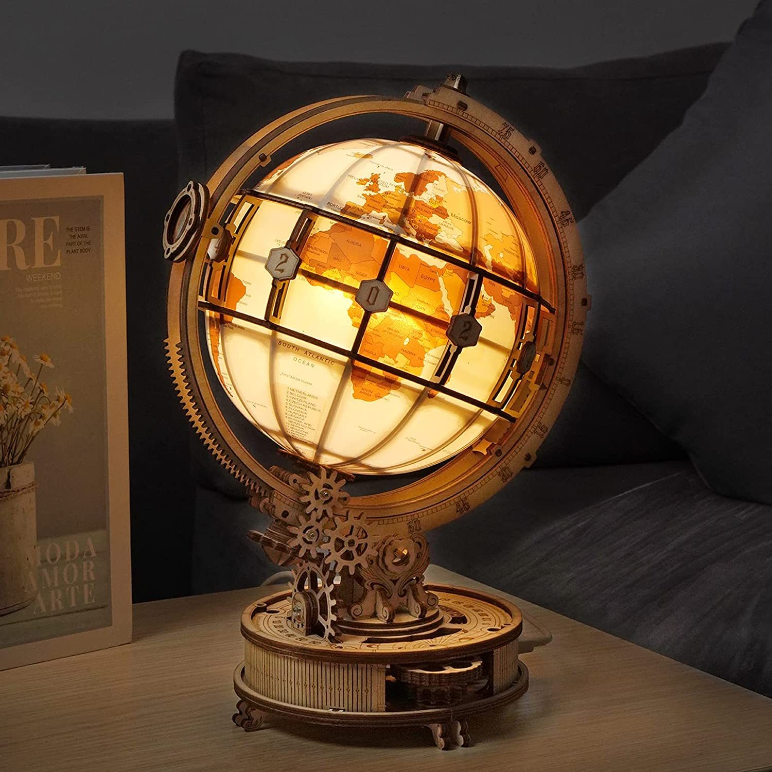 The image shows a wooden globe lamp sitting on top of a table next to a couch, with a book and a flower vase on the left side and a wall in the background. The lamp is made of wood and has a steampunk-style design, giving it a unique and eye-catching look.