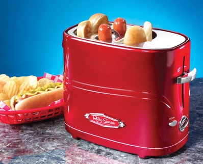 The image shows a red-colored toaster with a handle on the right side. The toaster has a label on the front with the brand name "Krispy Kreme" written in white letters. On top of the toaster, there are several hot dogs and buns, as well as a basket of french fries. The background is a blue-grey countertop.