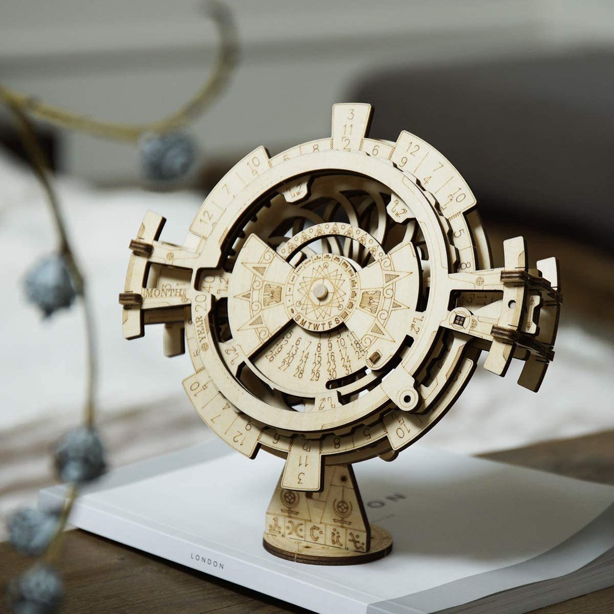 D rendering of a wooden model of a clock. The clock is circular in shape and appears to be made of a light-colored wood. It has a circular base with a pedestal at the bottom, which is also made of wood. The top of the clock has a clock face with numbers and symbols arranged in a symmetrical pattern. The numbers on the clock face are numbered from 1 to 12, and the symbols include a star, a crescent moon, and a starburst. There are also two arms on either side of the base, which are used to hold the clock in place. The arms are connected by a series of small holes, which could be screws or bolts. The overall design is intricate and detailed, with a vintage or antique feel.