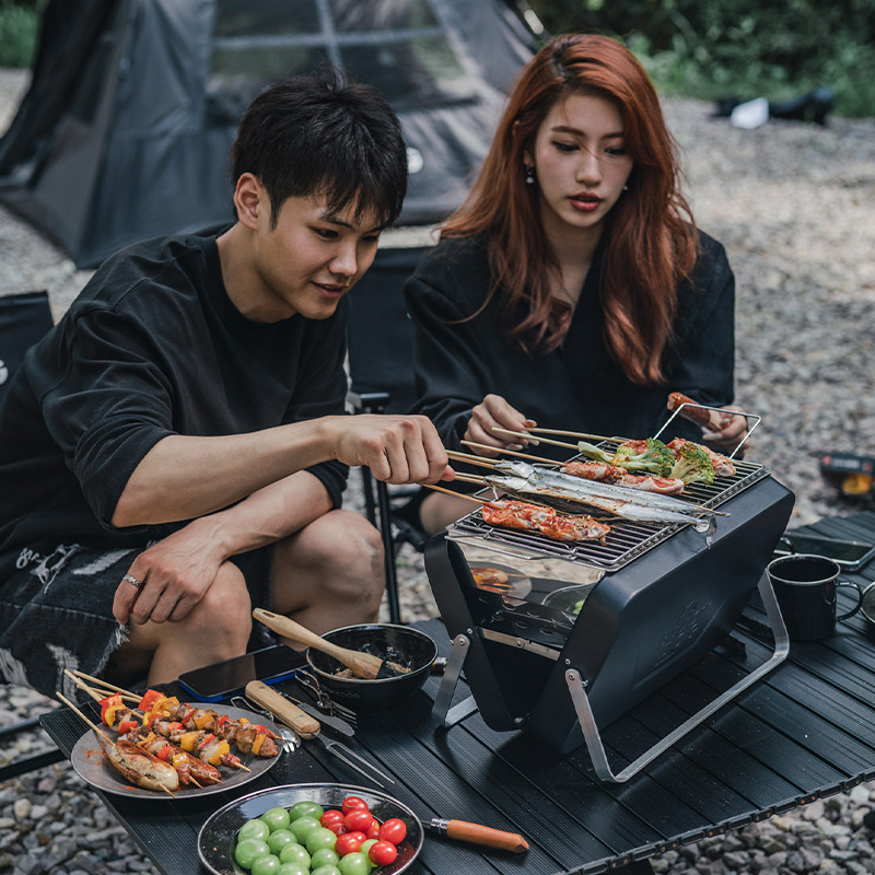 Photorealistic image of a young couple, a man and a woman, sitting on camping chairs at a low table, grilling skewers of food on a small portable charcoal grill. The woman has long reddish-brown hair, and the man has short dark hair. They are both smiling and engaged in the activity. The setting is outdoors, in a natural environment with greenery in the background. The scene is lit by natural daylight, with soft shadows. The overall mood is relaxed and convivial.