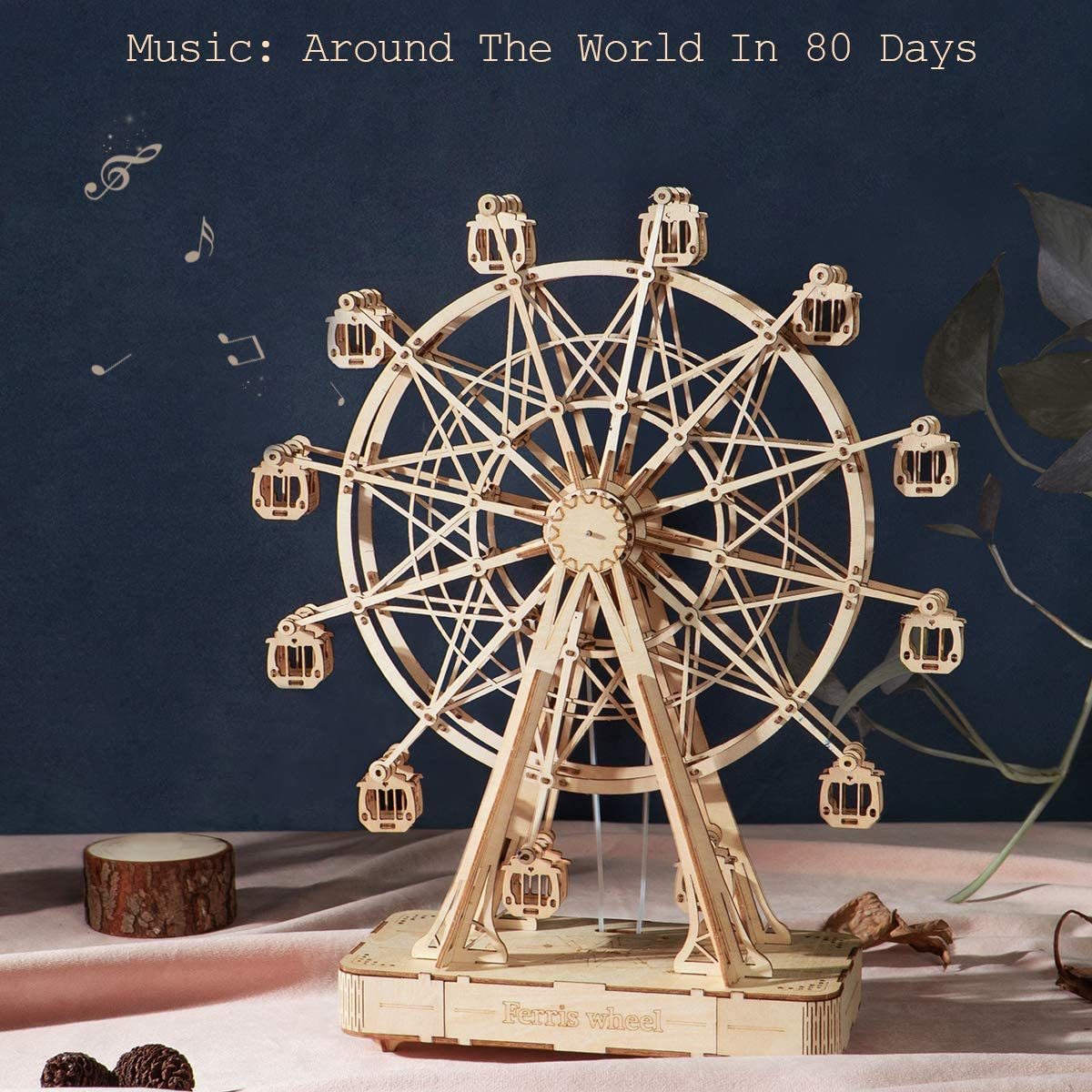 The image shows a wooden model of a ferris wheel on a table with the words "Music Around the World in 80 Days" written on it. The wheel is surrounded by a wooden object and other objects, and there is a wall in the background. At the bottom of the image, there are watermarks.