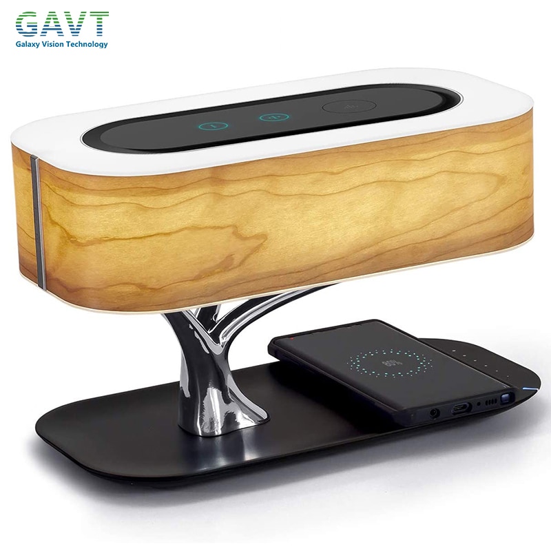 The image shows a wooden speaker with a unique design. The speaker has a rectangular shape with a flat top and a curved bottom. The wood has a light brown color with a natural grain pattern. It is supported by a silver metal base with a curved design. On the base, there is a black wireless charging pad with a circular logo on it. The charging pad is placed on a black base. The background is white.