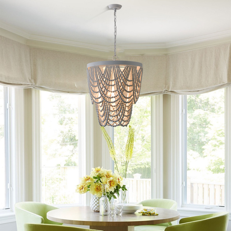 The image shows a dining room with a round wooden table in the center. The table is surrounded by four green chairs with a vase of yellow flowers on top. Above the table, there is a chandelier with a unique design made of woven baskets hanging from the ceiling. The room has large windows with white curtains, allowing natural light to enter the space. Through the windows, we can see trees and a wooden fence outside. The overall color scheme of the room is bright and airy.