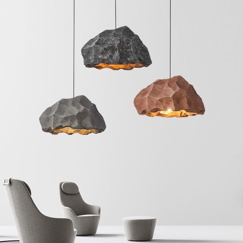 The image shows three pendant lights hanging from the ceiling in a modern living room. The lights are made of different materials, including a large rock-like object in the center, a smaller rock on the left, and a smaller one on the right. All three lights have a unique design, with the largest rock being the largest and the two smaller ones being the smallest. In the foreground, there is a gray armchair and a small white stool. The armchair has a curved backrest and armrests, while the stool has a round shape and a flat seat. The background is a plain white wall. The overall aesthetic of the room is minimalistic and contemporary.