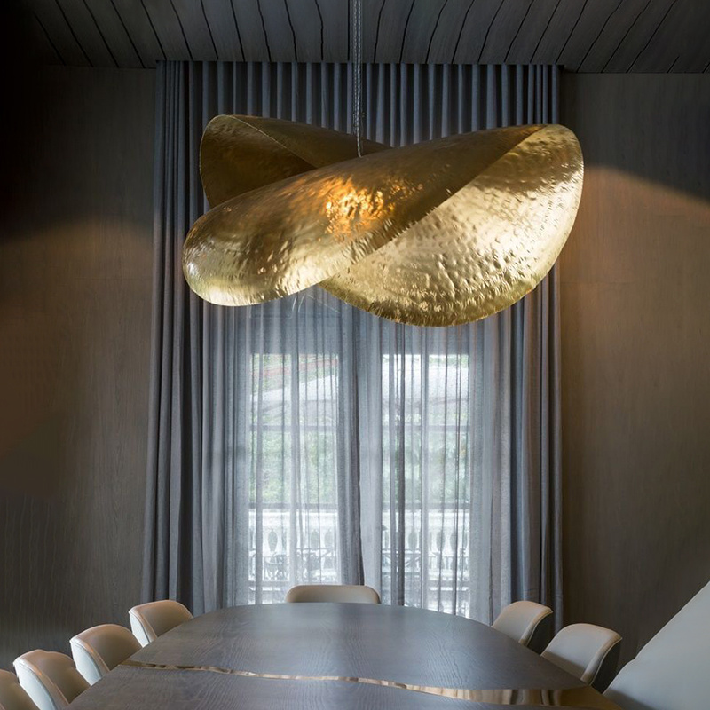 The image shows a modern dining room with a large wooden table in the center. The table is surrounded by white chairs and there is a large window with gray curtains in the background. Above the table, there are two large, gold-colored pendant lights hanging from the ceiling. The lights have a unique design with a curved shape and intricate details. The walls are painted in a dark grey color and the ceiling has a wooden paneling. The overall aesthetic of the room is elegant and sophisticated.