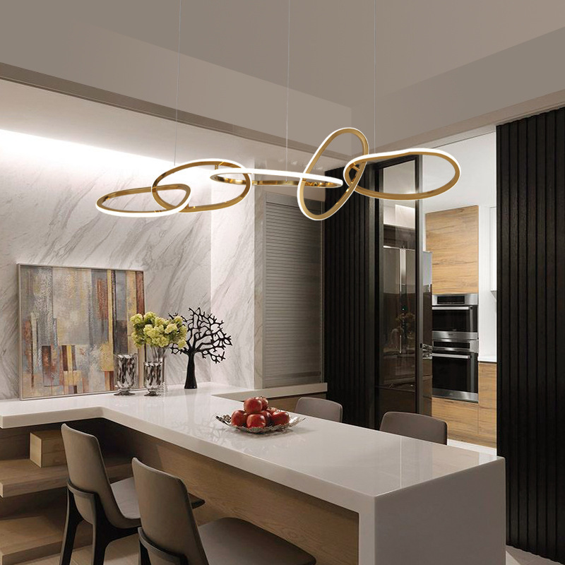 The image shows a modern kitchen with a large island in the center. The island has a marble countertop and a sink with a faucet. Above the island, there is a rectangular light fixture with a gold finish. The light fixture is made up of three curved lines that form an infinity symbol. The lines are connected by thin gold wires, creating a unique and eye-catching design. The overall color scheme of the kitchen is gray and white, with a touch of gold accents.