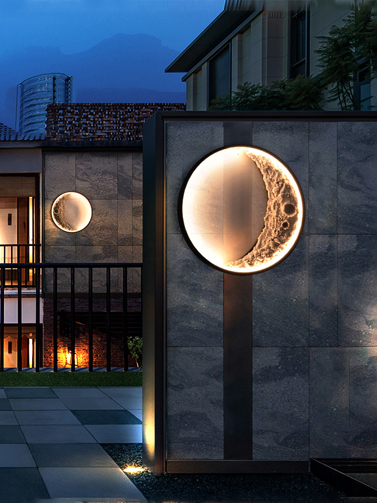 The image shows a modern building with a large circular light fixture on the side of it. The light fixture is made of metal and is mounted on a gray concrete wall. The wall is illuminated with a warm orange glow, creating a contrast against the dark background. In the background, there is a view of the city skyline and a mountain range. The building appears to be a two-story structure with a balcony and a balcony railing. The sky is dark, suggesting that the photo was taken at night.