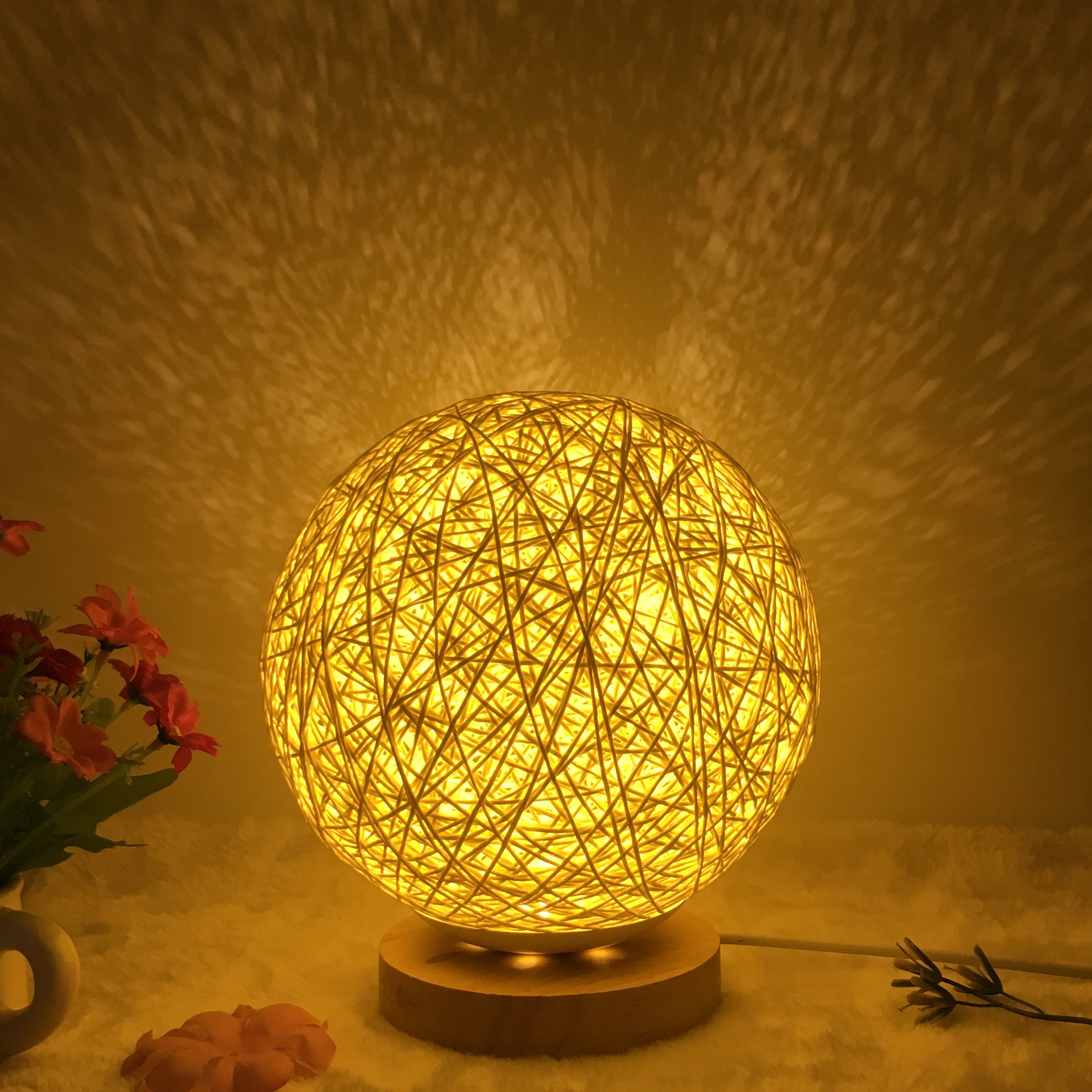 Warm glowing Creative Rattan Ball Lamp with hand-knitted shade on wooden stand, casting intricate shadows on the wall.