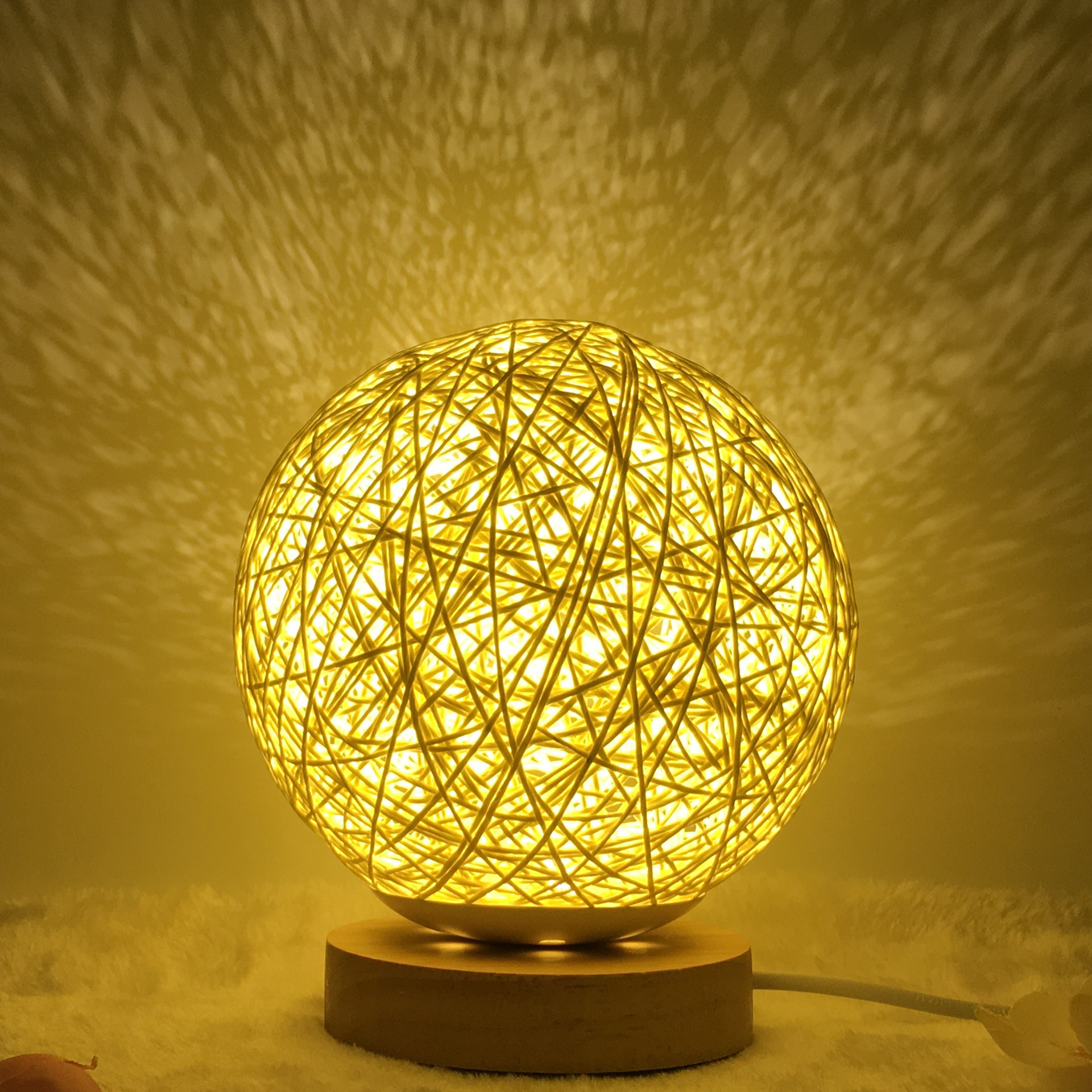Warm glowing Creative Rattan Ball Lamp with hand-knitted shade on wooden stand, casting intricate shadows on the wall.