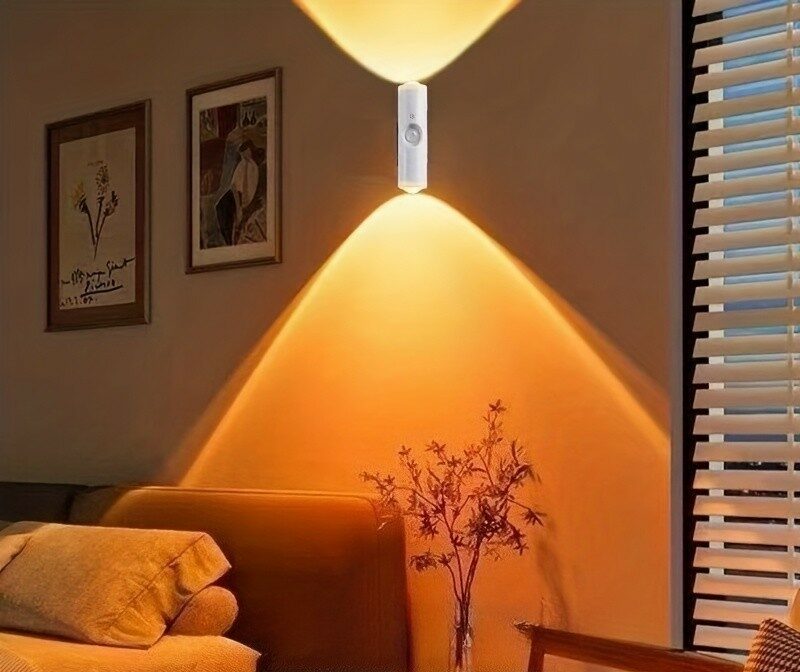The Aura Magnetic Motion Sconce White cylindrical wall light casting warm amber beams up and down a wall beside a sofa and framed pictures